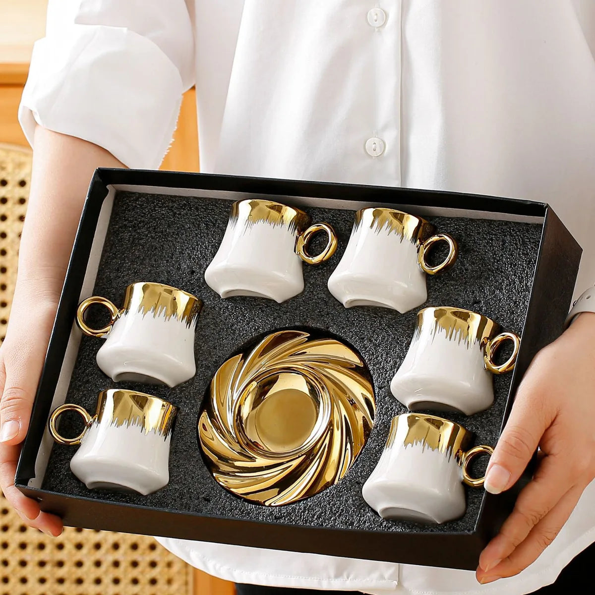 Set of white and gold ceramic cups and saucers in a black box held by a person wearing a white shirt.