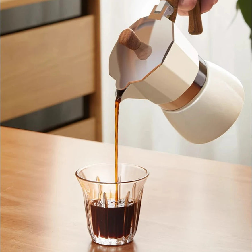 Person pouring coffee from a white and wood coffee pot into a glass on a wooden table.