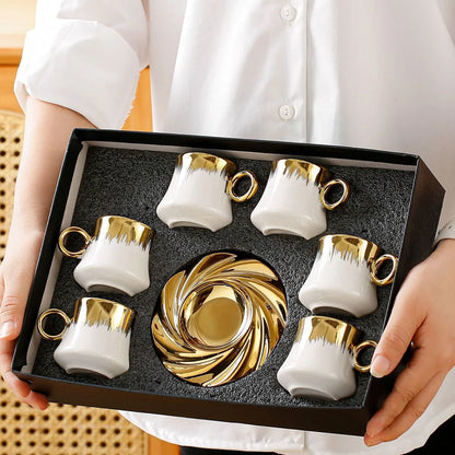 Set of white and gold ceramic cups and saucers in a black box held by a person wearing a white shirt.