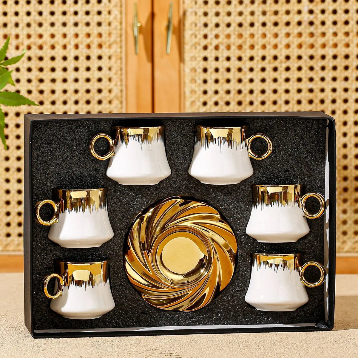 Set of white and gold ceramic cups with a decorative plate in a black box against a woven background.