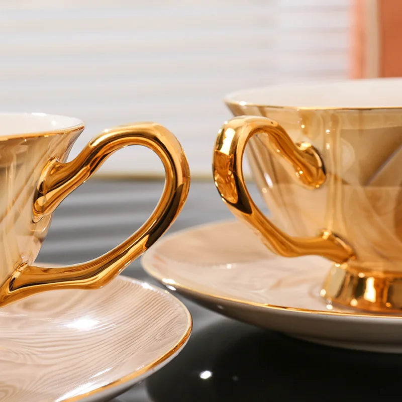 Two gold-rimmed coffee cups and saucers on a reflective surface with a blurred background.